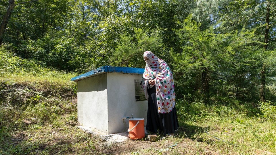 A woman stands at a small cement building, filling her cooler with water from a faucet