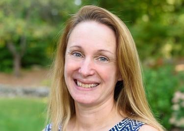 A headshot of Tara Happy who has long yellow hair and wears a blue patterened top and smiles at the camera in front of a green outdoor background