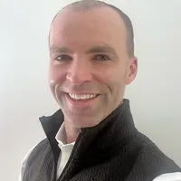 John Ambler wears a black vest and white shirt and smiles at the camera in front of an