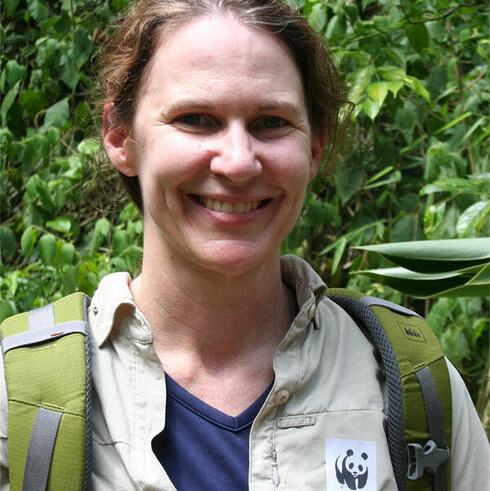 A headshot of Linda Walker wearing a tan shirt and green backpack smiling at the camera with greenery in the background