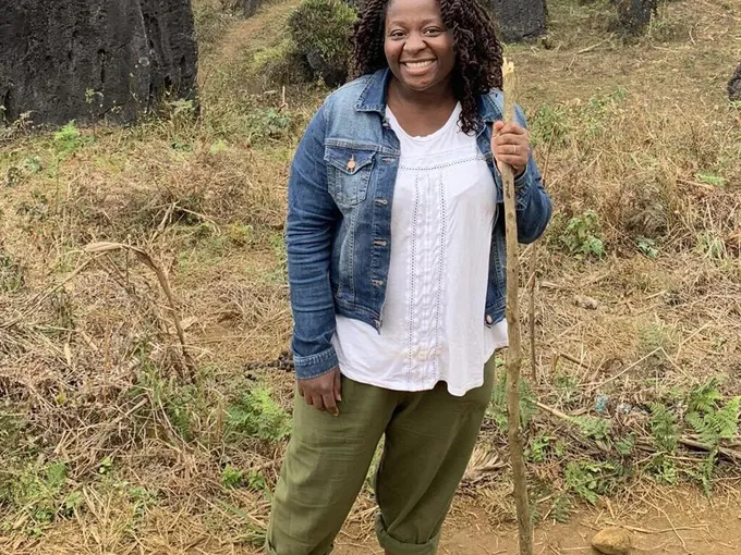 Florence Adewale stands on a trail with a hiking stick in a blue jacket