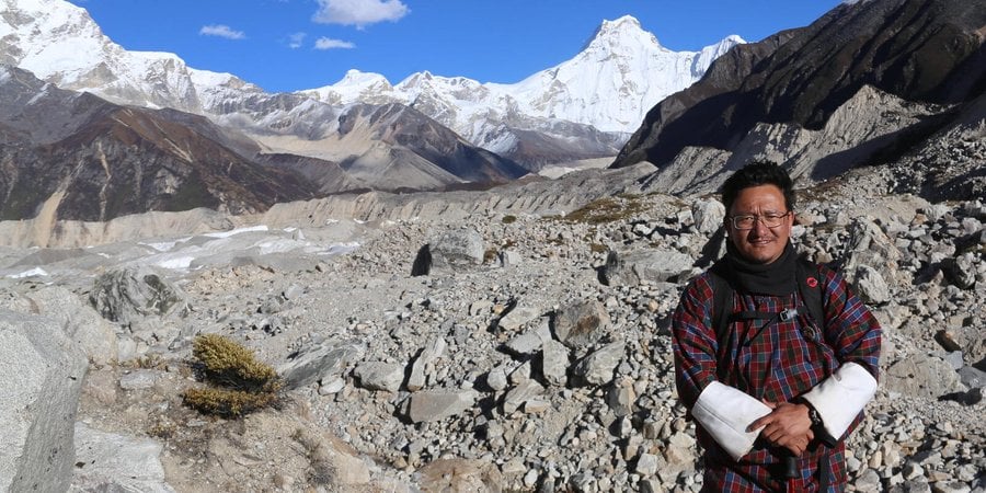 Dechen Dorji stands in front of tall snowy peaks on a sunny day