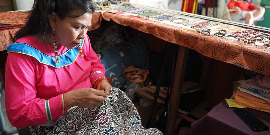 A woman wearing pink stitches a patterned fabric next to a table full of beaded jewelry.