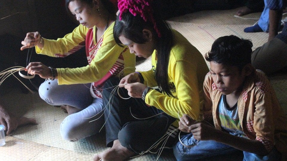 Three women sit on the floor weaving a mat out of straw