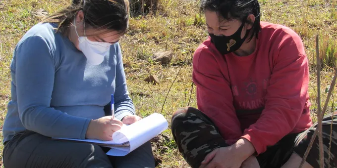 Two women sit on the grassy ground, one holds a notepad taking notes and each wear face masks
