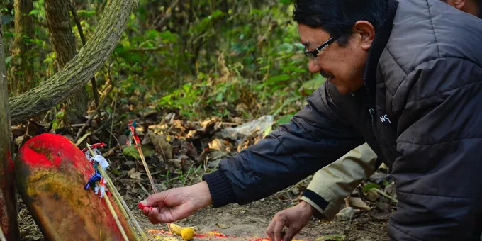 A man kneels down on the ground in the forest to place red powder on a stone