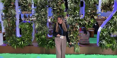 Woman stands in front of a leafy green backdrop smiling at the camera and holding a badge that's around her neck