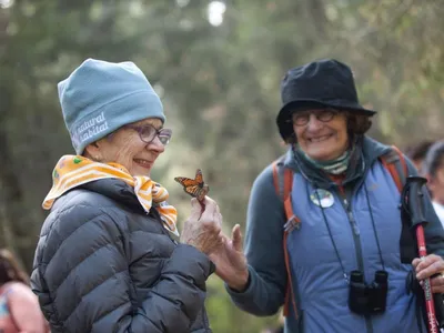 Two women smiling widely at a monarch butterfly that has handed on one of their hands