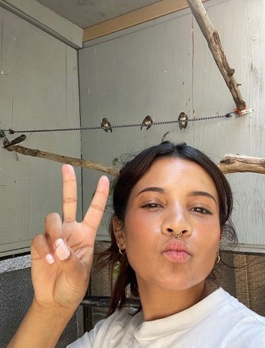 Nikki Rodriguez has dark hair and wears a white shirt. She is making a peace sign.