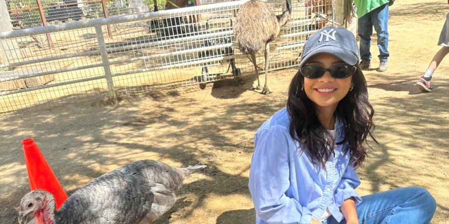 Nikki Rodriguez sits on the ground wearing all blue, a hat, and sunglasses, next to a bird