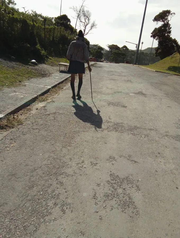 A man in a hat, with a basket and walking stick, walks away down a road