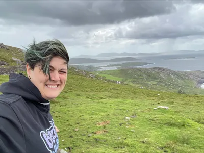 A picture of Sam-Mayes Matthews looking at the camera from a green coastline under a cloudy sky