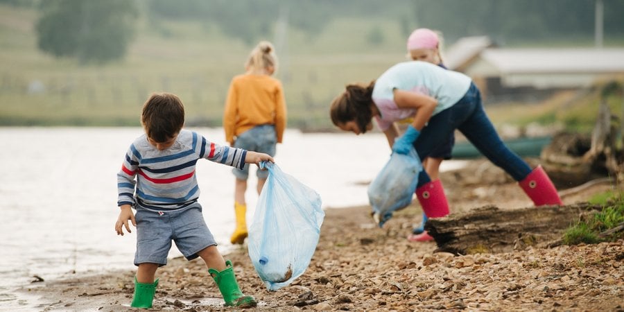 A young boy in a wearing rain boots and a plastic garbage looks down as he collects trash along a shoreline. Other children can be seen behind him doing the same thing.