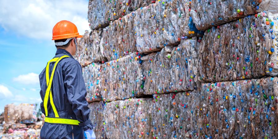 A person in a blue jumpsuit and orange hardhat stands in front of a wall of plastics being recycled