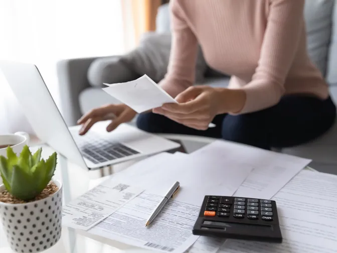 Close up crop of a woman working on laptop at home with paper and a calculator.
