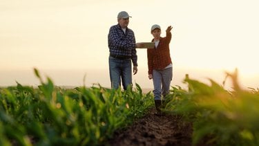 Two farmers stand in a field. One holds a digital tablet and the other farmer points to field with hand.