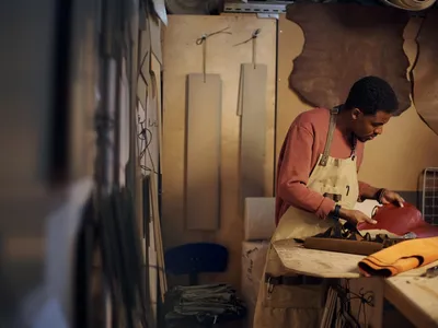 Young artisan meticulously crafting leather good in warmly lit workshop. Table is covered with tools and leather pieces