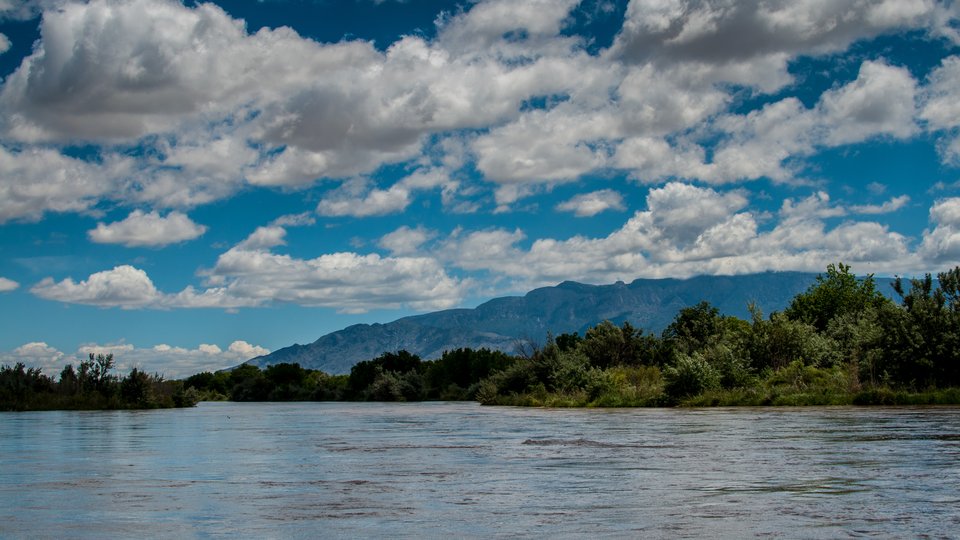 A color photo showing the Rio Grande River in the foreground, trees and mountains in the background with a blue sky filled with white clouds