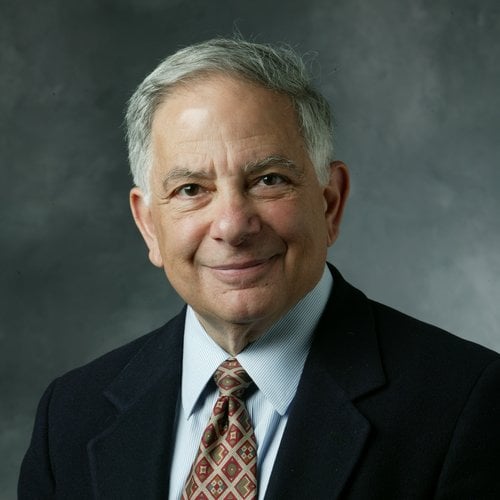 Headshot of a smiling white-haired man. He is wearing a black suit and a red patterned tie.