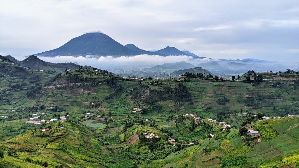 Rolling green hills with homes on them and large cloud-covered mountains in the background