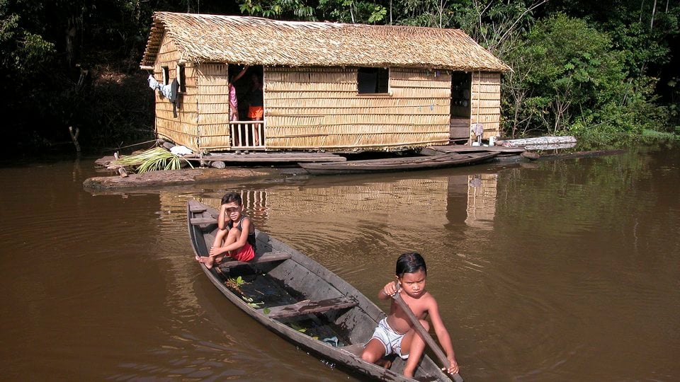 Two small children in a wooden kayak in the foreground and a murky brown river and wooden house in the background.