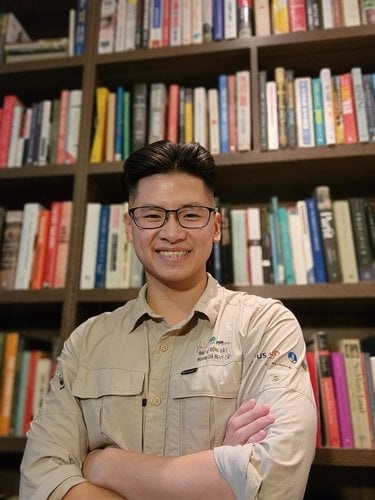 An Asian person with short hair and an under cut smiles, has black glasses, and wears a tan, long sleeve button down.