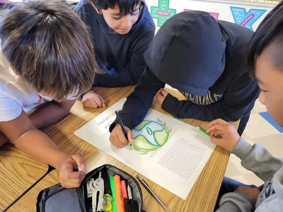Students sit at a table watching another student color in a coloring page of Earth