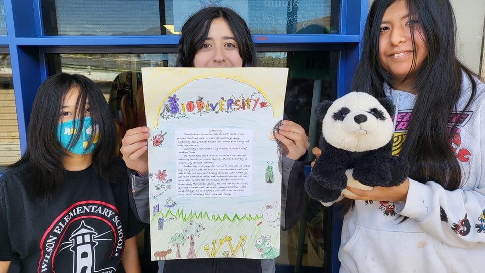 The photo is of three students. One is holding up a hand-drawn poster about biodiversity and another is holding a panda plush.