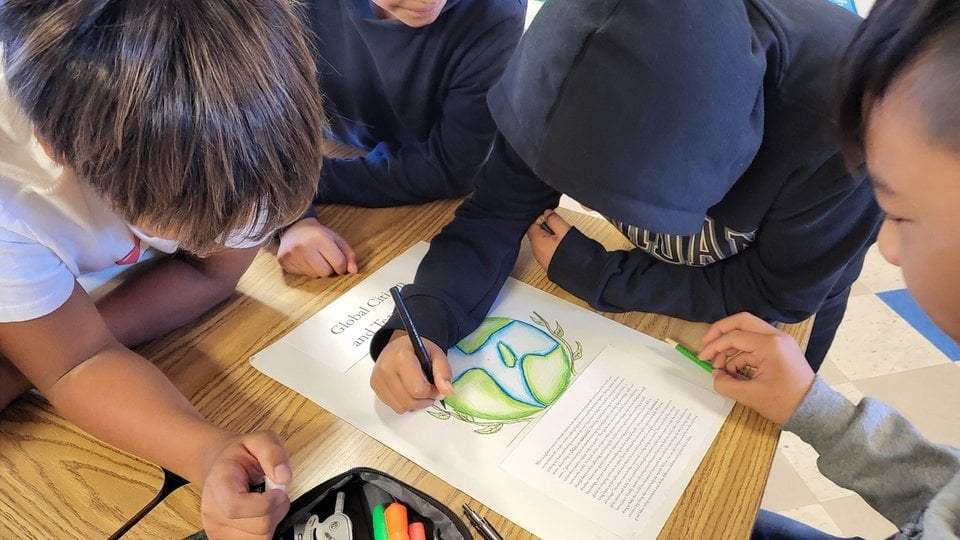 Students sit at a table watching another student color in a coloring page of Earth