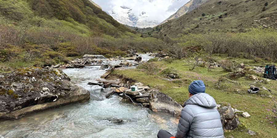 A person in a puffy gray jacket sits on a rock by a mountain stream while a pump collects water samples nearby.