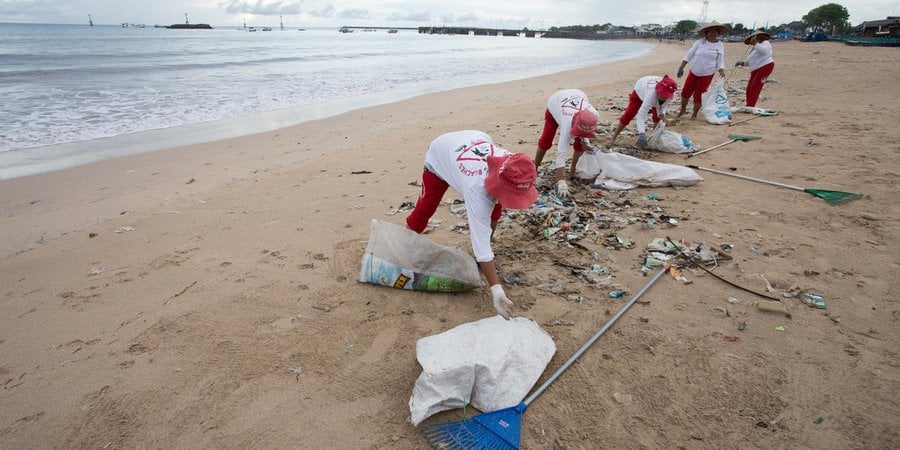People in white shirts and red hats pick up plastic waste on a beach in Indonesia.