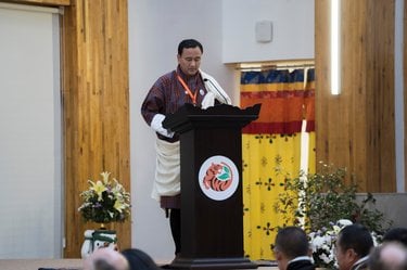 A man in traditional clothing stands behind a podium, speaking to a crowd