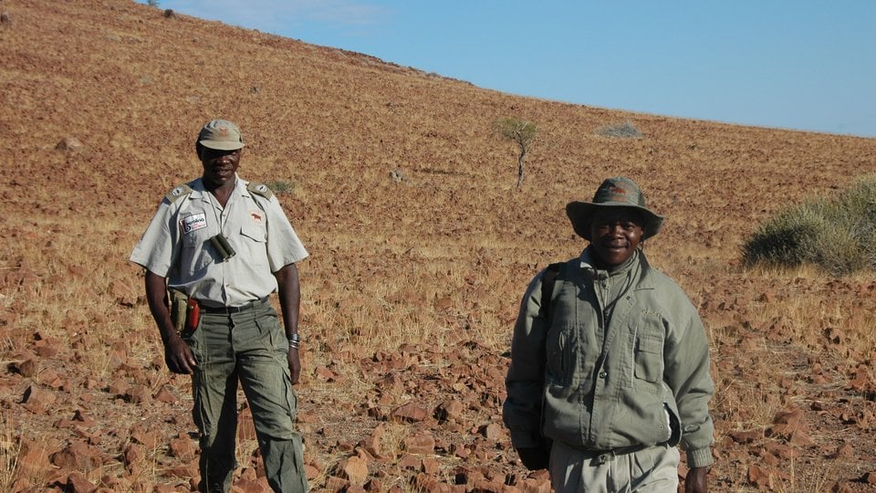 Two men in cargo clothing stand in an expansive rocky yellow grassland