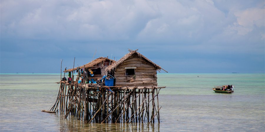 Small woven houses sitting on top of a system of stilts out in clear blue water with a green boat anchored nearby