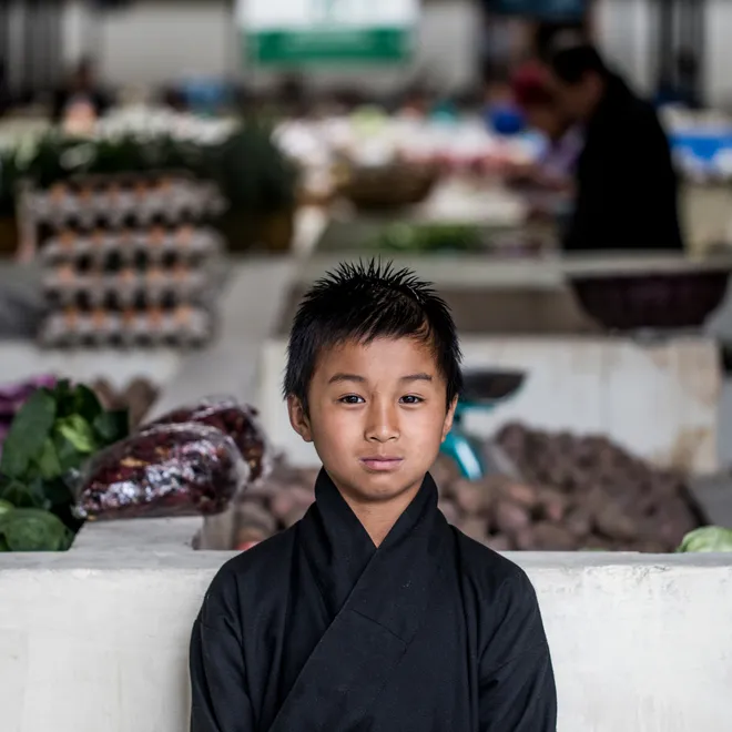 A Bhutanese boy wearing traditional clothing stands in front of a vegetable stall at a market.