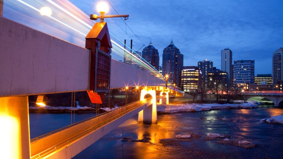 A night time view of a illuminated, white bridge in Calgary Alberta. It spans a river with snow along the edges and tall buildings of a city in the distance.