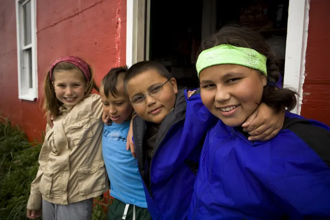 Four school kids from the Bristol Bay area stand in a line, hold shoulders and smile in front of a red house.