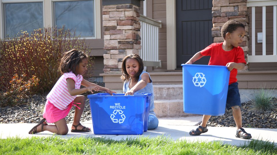 Children place items in a recycling bin in front of a house