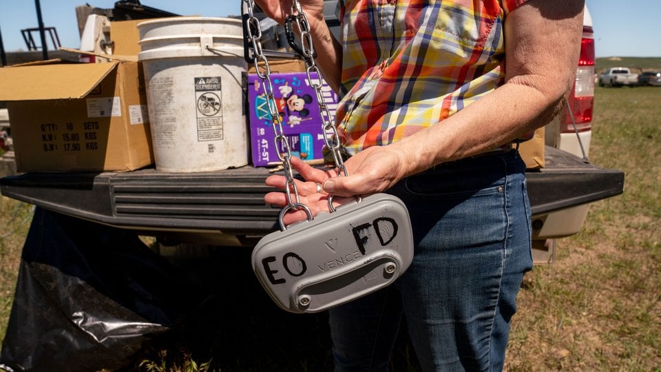 A person in a plaid shirt holds a gray box affixed to a chain that serves as a virtual fencing collar