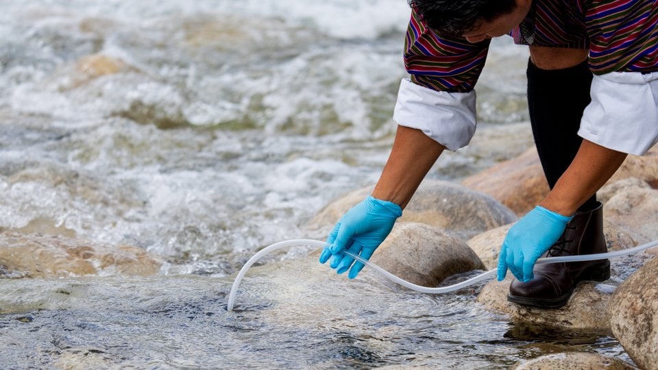 A close up of hands wearing blue gloves and holding a tube to collect water from a stream