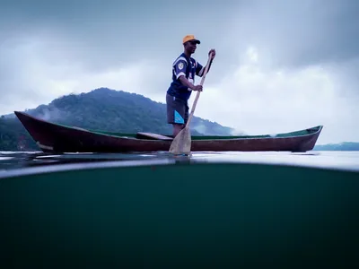 A man rides a small canoe in flat green water with a green mountain in the distance and gray clouds.