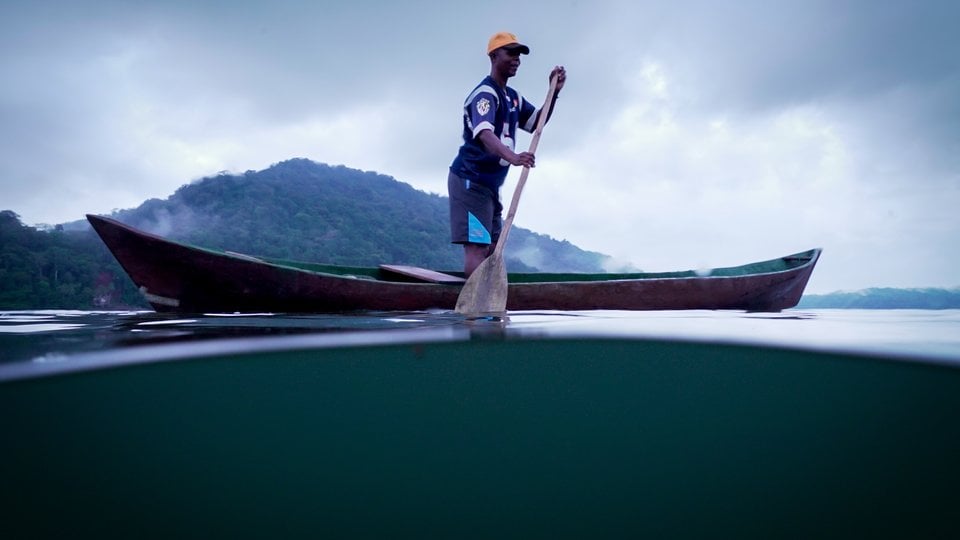 A man rides a small canoe in flat green water with a green mountain in the distance and gray clouds.