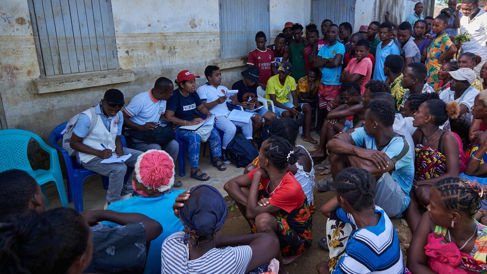 Large group of people meeting outside, some sitting in the shade of a building