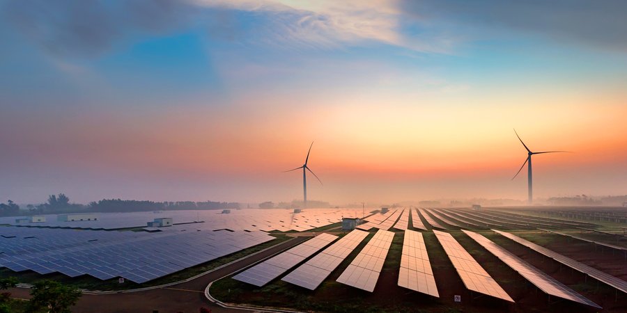Landscape photo of a field of solar panels at sunrise. There are two wind turbines in the distance. Everything is shrouded in a light mist.