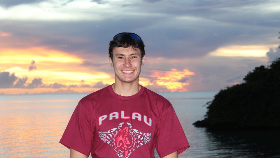 A color photo showing a man wearing a Palau tshirt in front of a sunset over water in the background