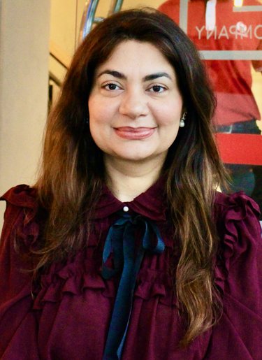 A headshot of Dr. Khan. She has long, wavy brown hair and is wearing a maroon shirt with black ribbon.
