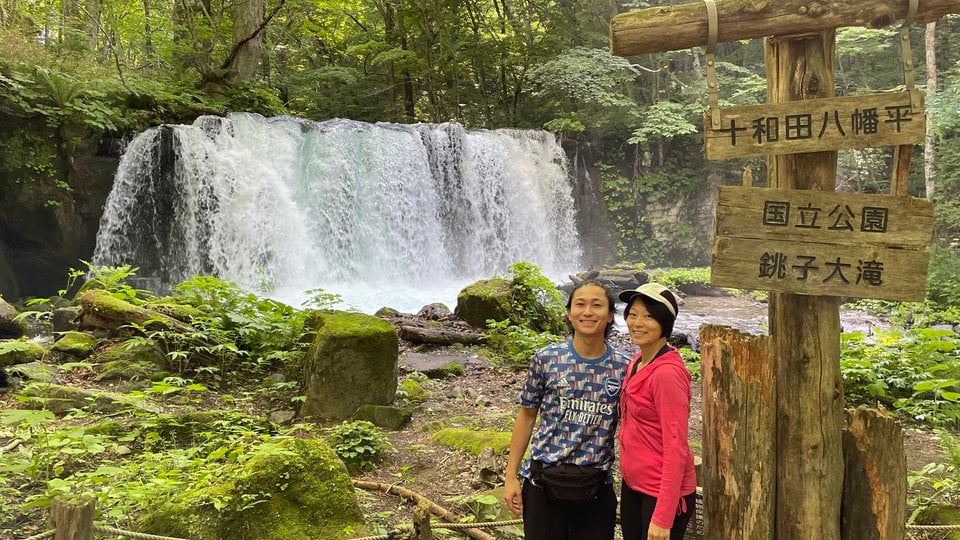 A color photo of two people standing in front of a waterfall