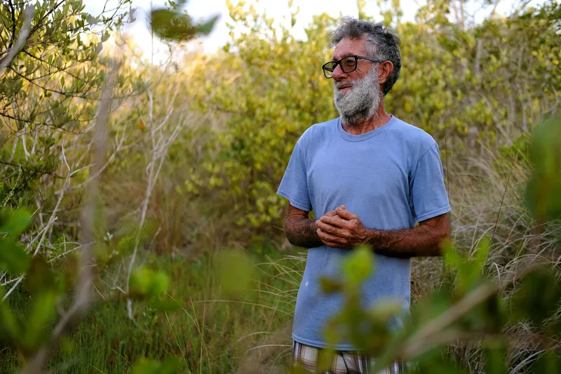 A bearded man with glasses and a light blue shirt standing surrounded by green shrubs.