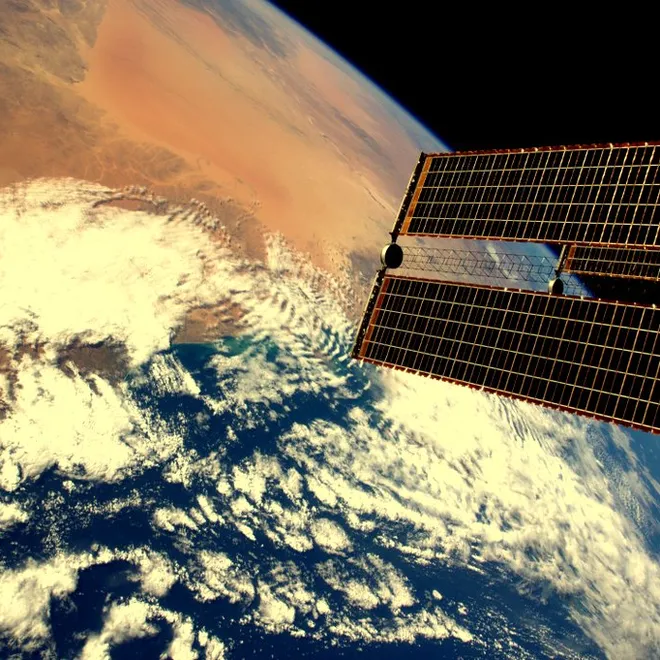 An image of Earth, taken from space that shows a desert climate on earth and has a satellites solar array in the foreground