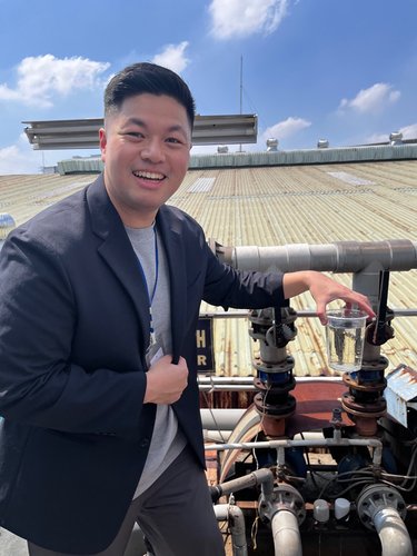 An Asian man wearing a blazer holds a cup of water and poses in front of machinery.